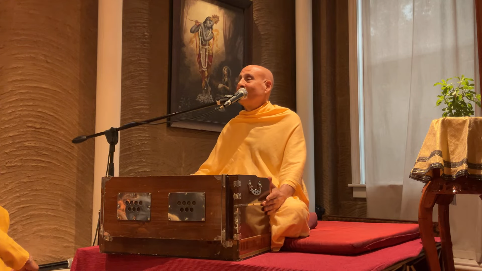 Bald man in saffron robes seated behind a harmonium and speaking or singing into a microphone.