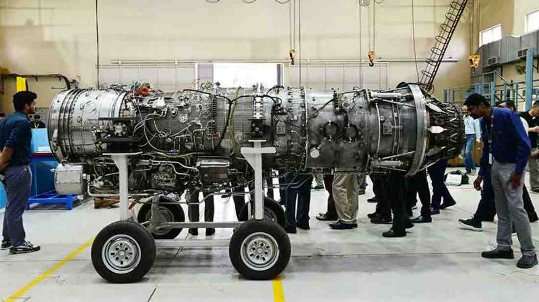 Large cylindrical engine assembly on a wheeled stand as several people inspect it in an industrial workshop.