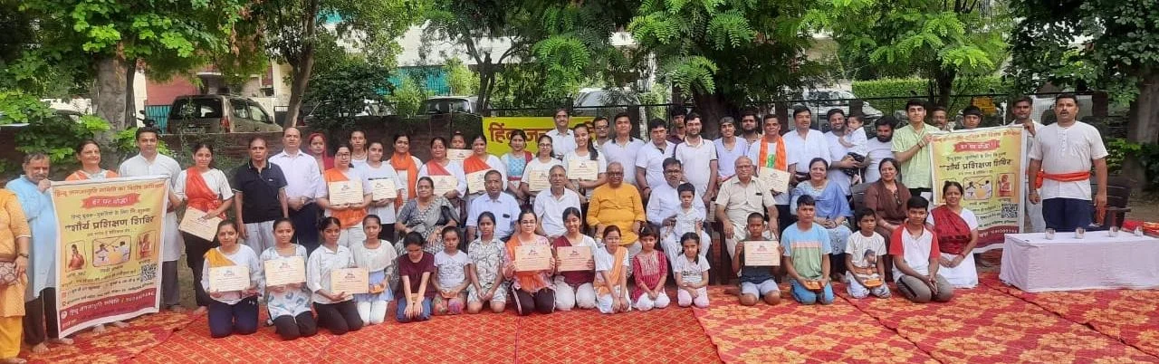 Large group of adults and children posing outdoors with certificates and two Hindi banners