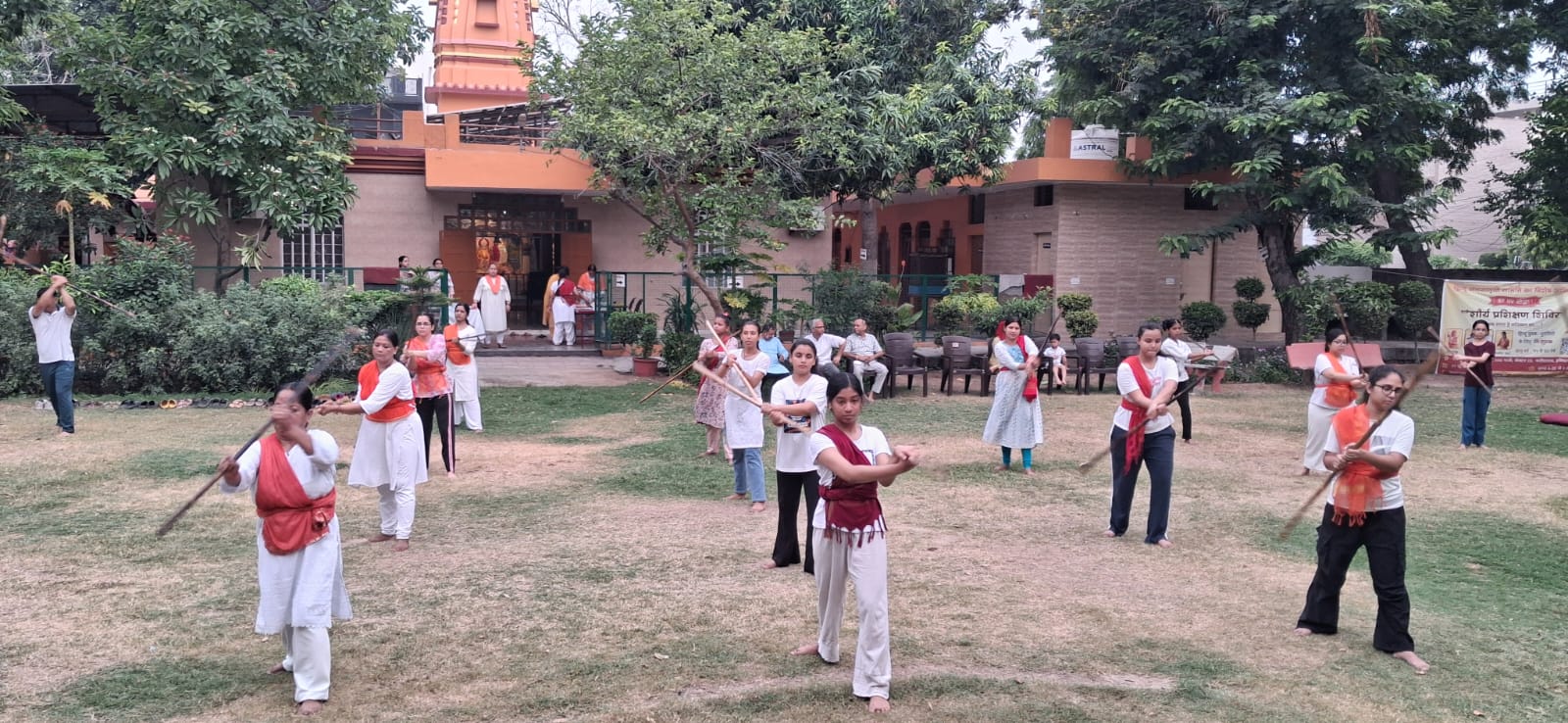 Women practice coordinated stick movements on a lawn while onlookers sit outside a tree-lined building.