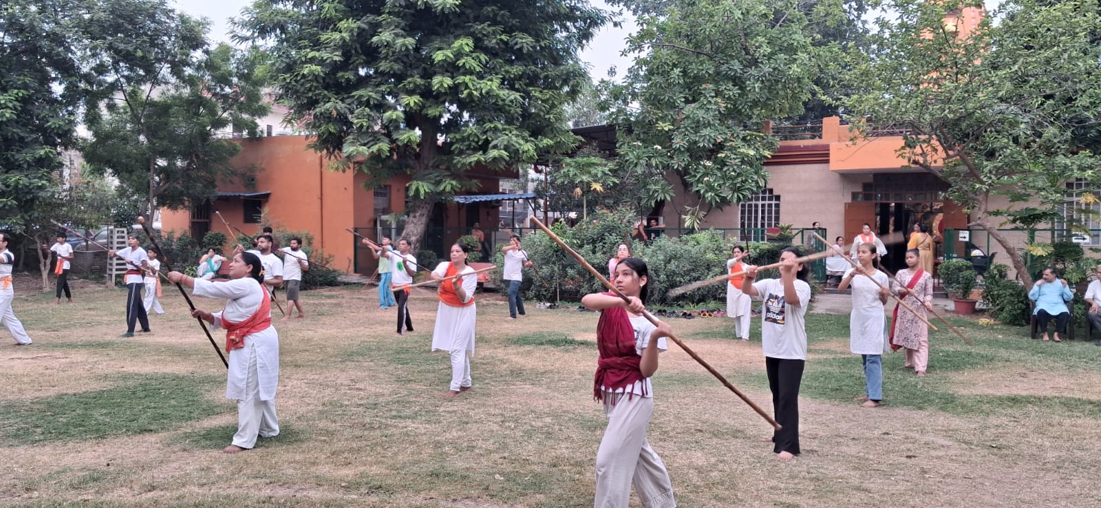 A group of people practice coordinated wooden-staff movements on a grassy lawn bordered by trees and buildings.
