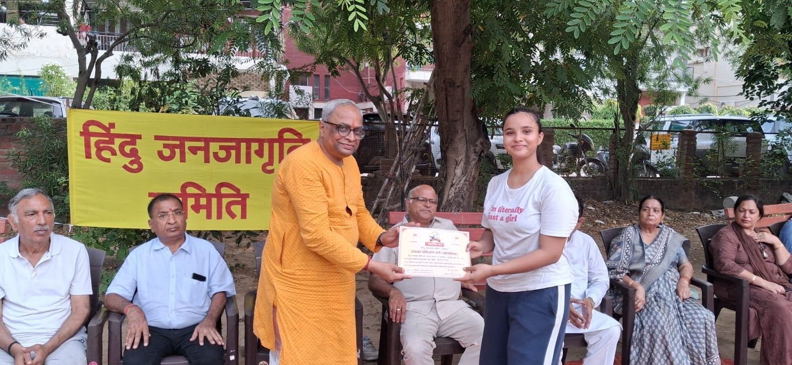 A man in an orange kurta presents a certificate to a young woman as seated adults look on beside a yellow Hindi banner.