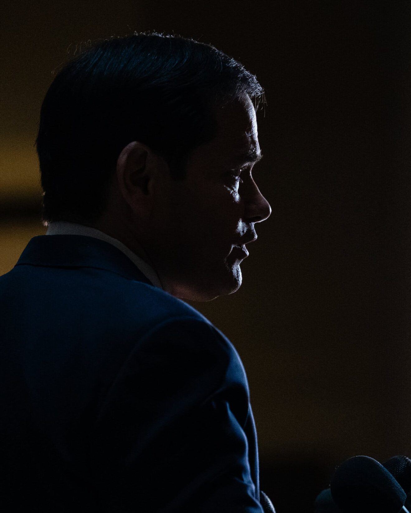 Dimly lit side profile of a man in a dark suit, with microphones visible at the lower right.