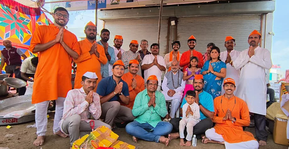 Group of adults and children in orange and white clothing posing with folded hands outside closed shop shutters.