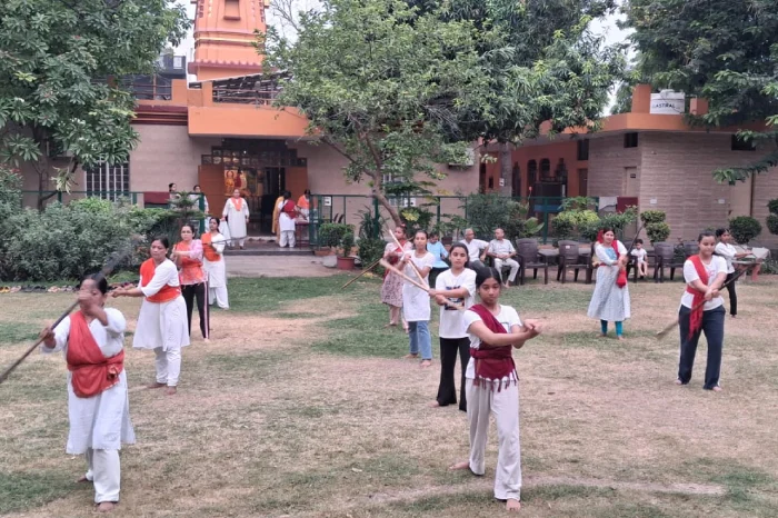 Women perform a coordinated stick routine on a lawn outside a building as onlookers sit nearby.