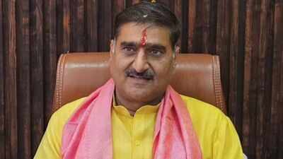Seated man in a yellow kurta and pink scarf, with a red tilak and moustache, against dark wood panelling.