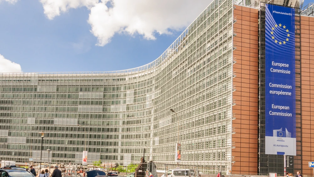 Curved glass office building beside a blue European Commission banner displaying the EU circle of stars.