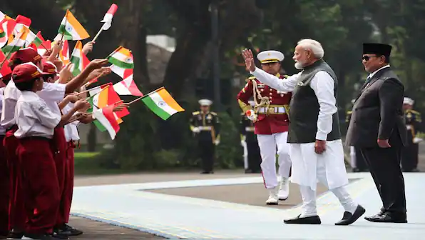 White-haired man in a dark vest waves to uniformed children holding Indian tricolor and red-and-white flags.