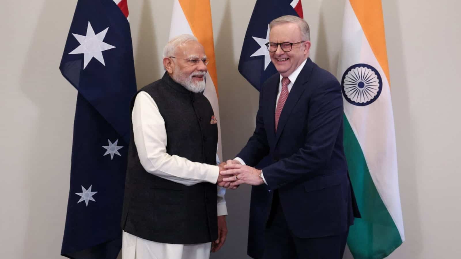 Two smiling men shake hands in front of alternating Australian and Indian flags.