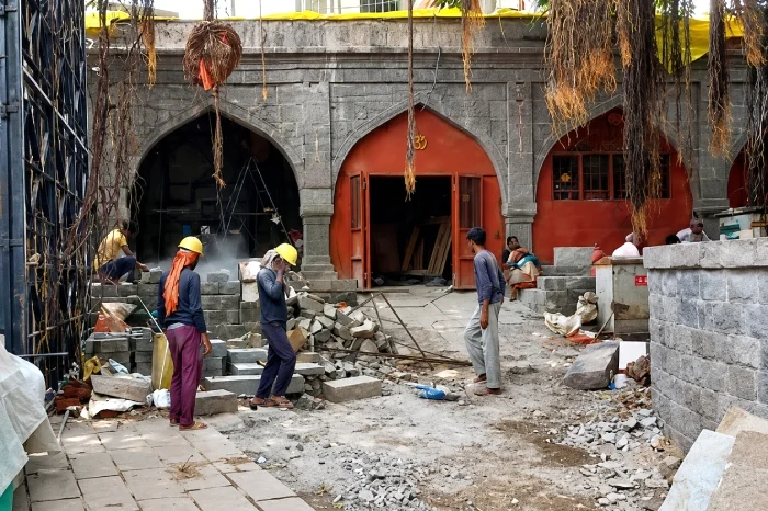 Workers in hard hats stand among stone blocks and rubble outside an arched gray stone building.