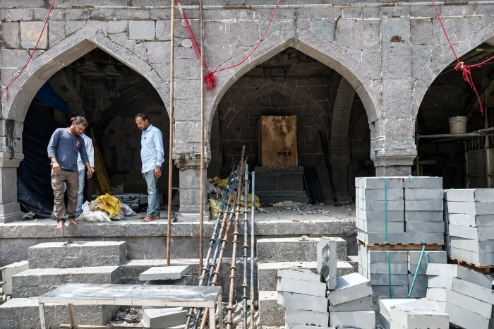 Two men stand inside a stone arcade amid stacked masonry blocks, pipes, steps and construction debris.