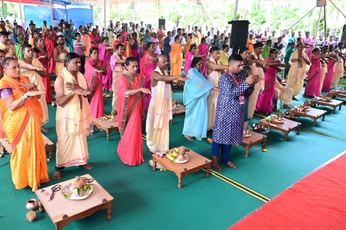 A large group in colorful traditional clothing performs synchronized gestures beside low tables of offerings.