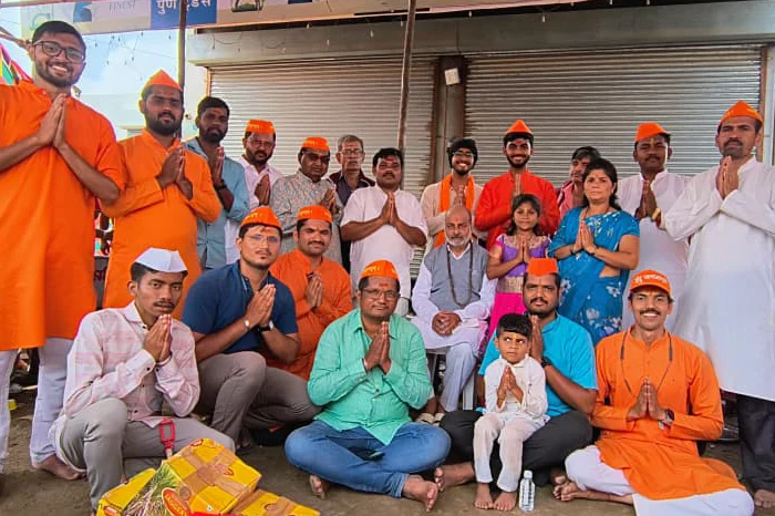 Group of adults and two children posing with folded hands, many wearing orange caps, in front of closed shutters.