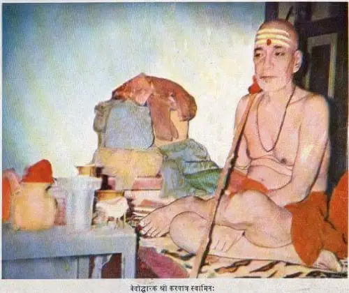 Elderly ascetic seated cross-legged indoors, holding a long staff beside vessels, cloth bundles, and bedding.