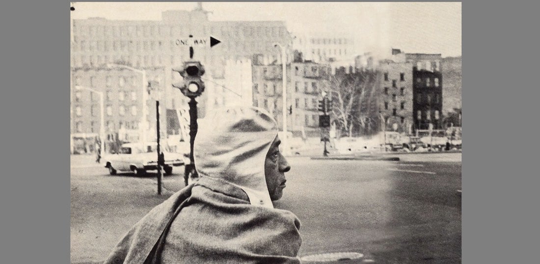 Hooded person wrapped in a shawl looks across a city intersection in a grainy black-and-white photograph.