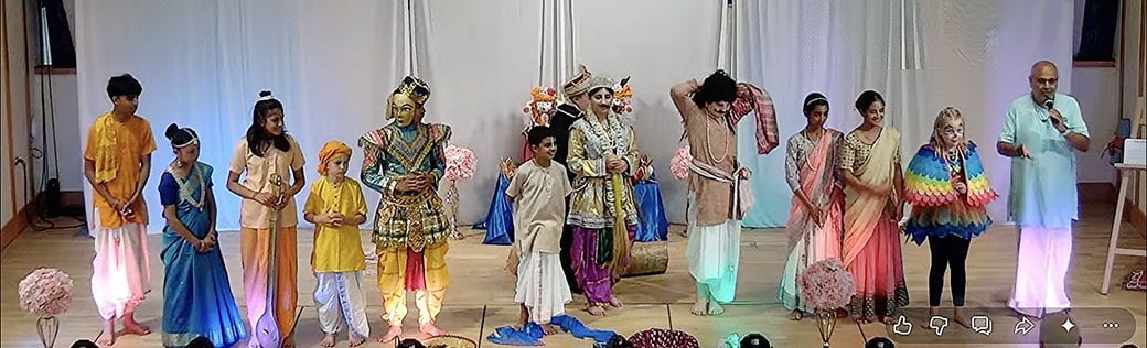 Children and adults in colorful costumes line a stage beside a man speaking into a microphone.