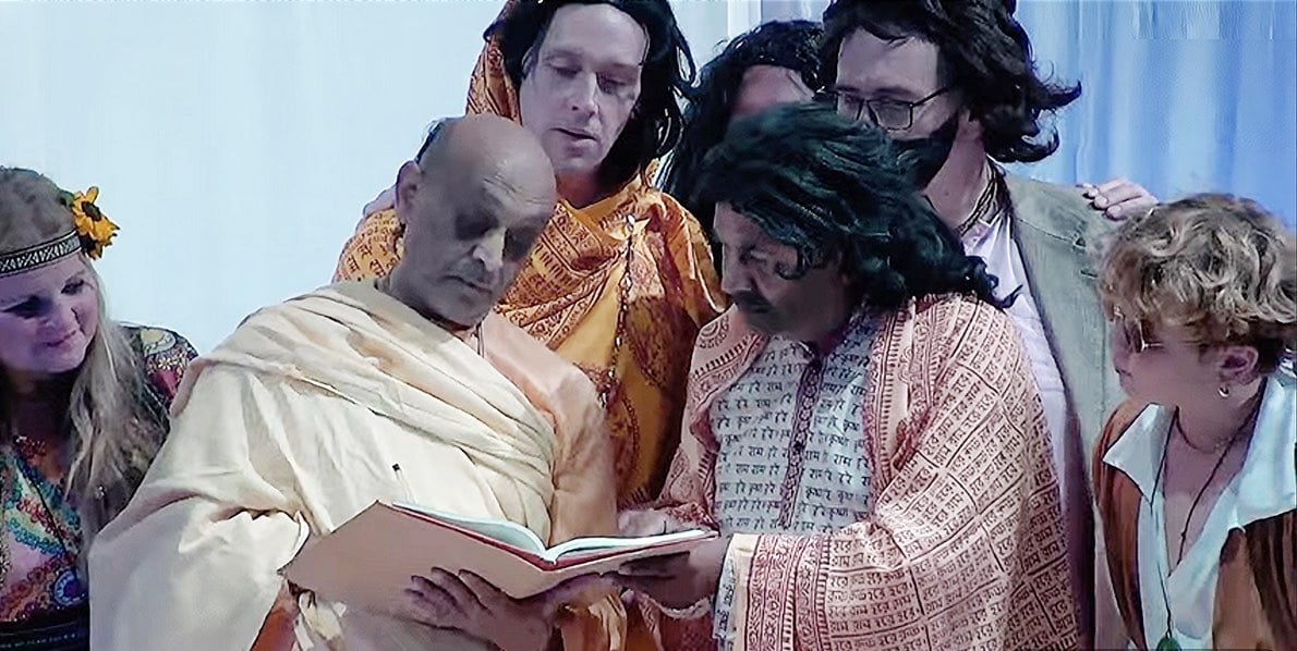 Seven adults in theatrical dress gather around an open book held by two robed figures.