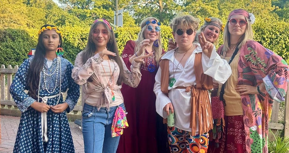 Six people in colorful retro outfits pose outdoors with sunglasses, flower crowns and peace-sign necklaces.