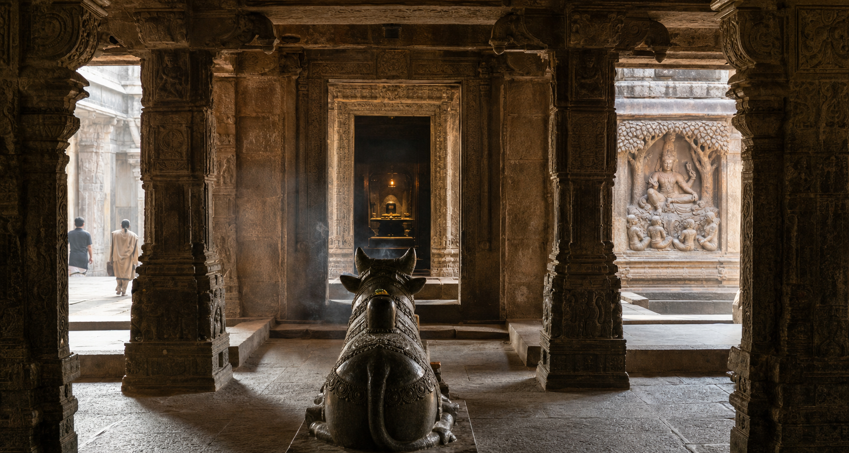 Nandi faces a lamp-lit Shiva linga along the central axis of a pillared stone temple, while a Dakshinamurti carving occupies a side niche on the sanctum wall.