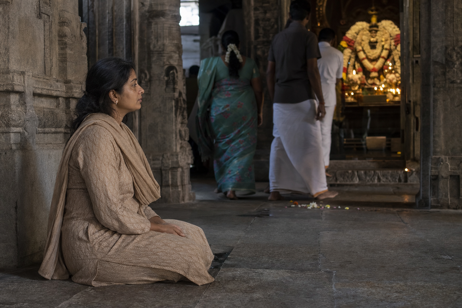 A seated devotee rests calmly near a temple sanctum while keeping her eyes directed toward the distant deity murti.