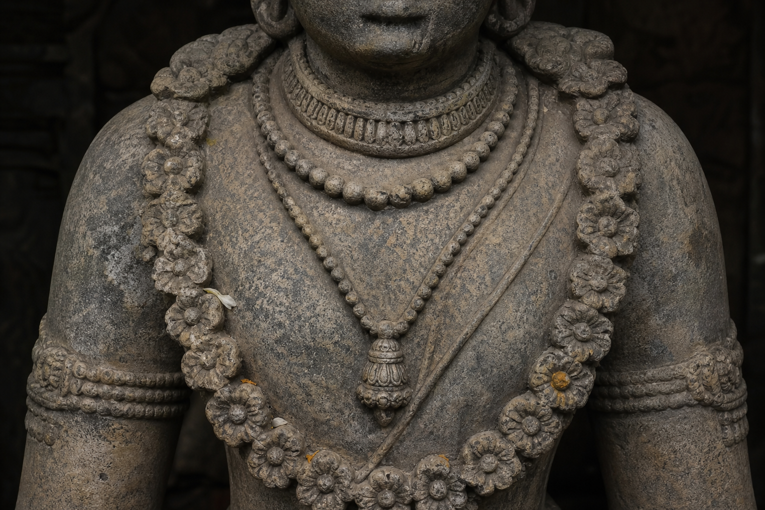 Close view of a stone murti's upper torso showing a collar, beaded necklaces, a long garland and a diagonal sacred cord as distinct carved elements.