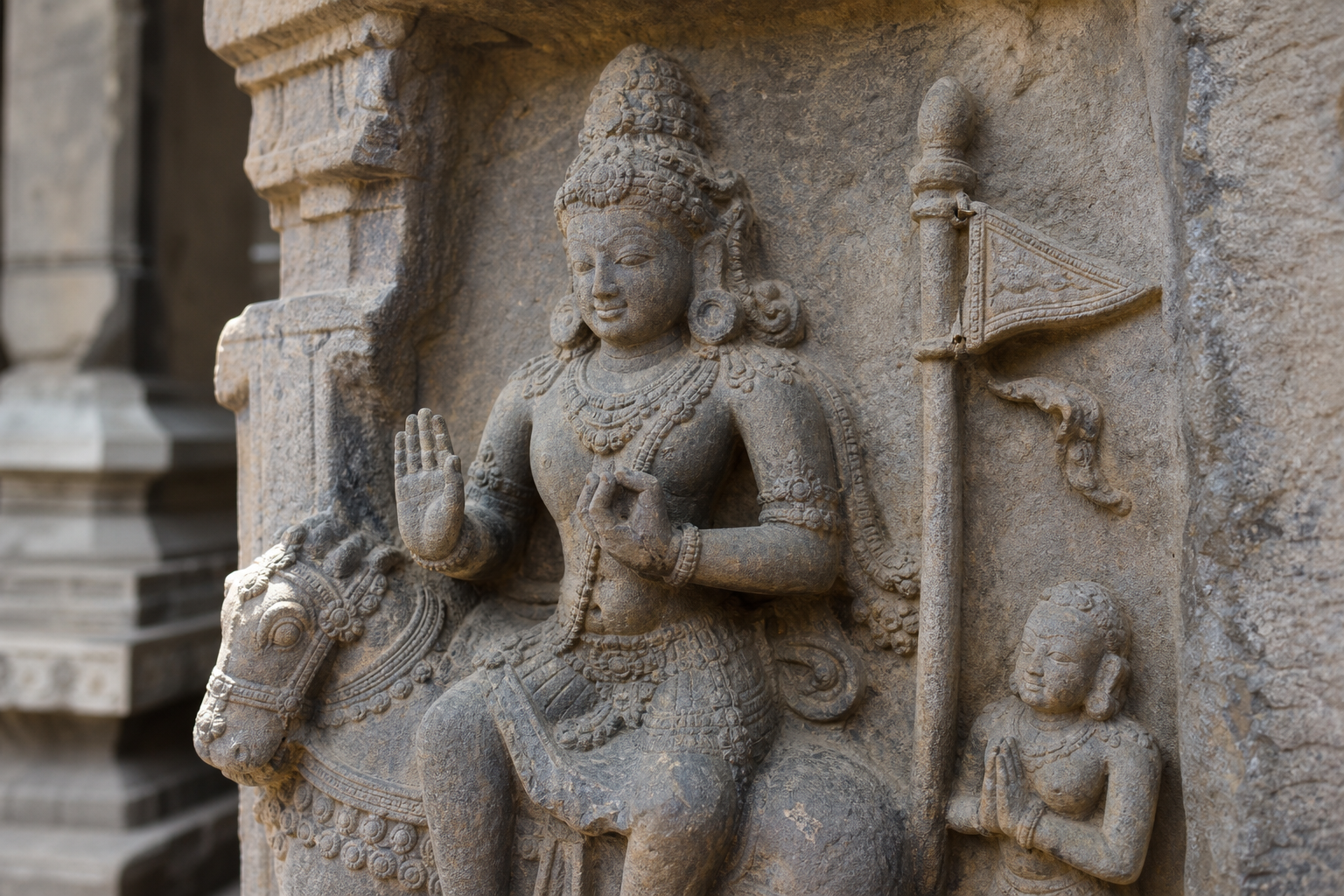 Weathered stone relief of a divine figure with a small pennon positioned beside the figure among other carved attributes.