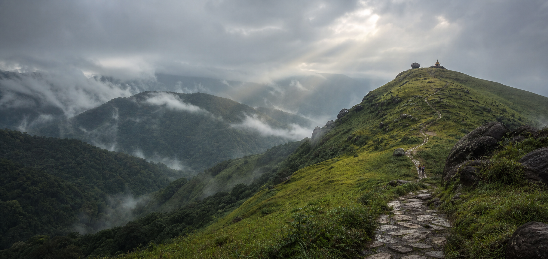 Pilgrims follow a stone path across misty green ridges toward a small hilltop shrine in the Chikkamagaluru hills.