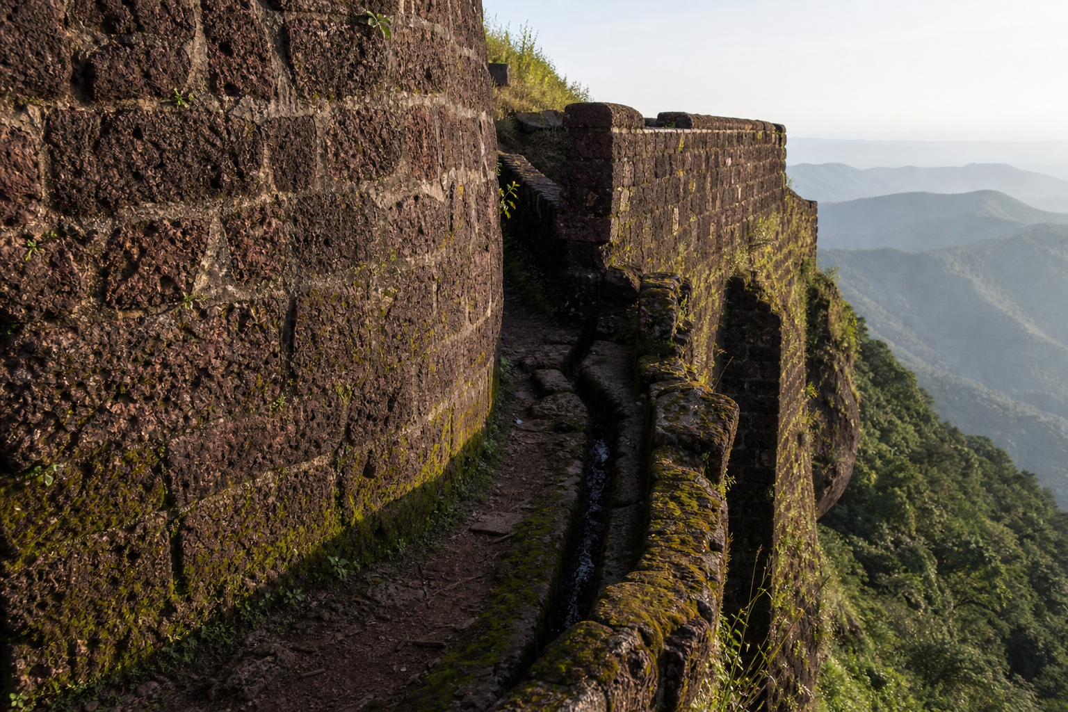 An eroded laterite bastion and defensive wall at Vishalgad stand beside a narrow path above the green Sahyadri escarpment.