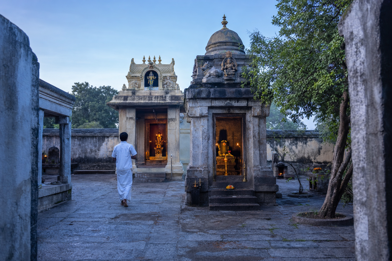 A pilgrim approaches a modest South Indian temple complex at dawn, with the Shiva shrine in front of an illuminated Murugan sanctum.