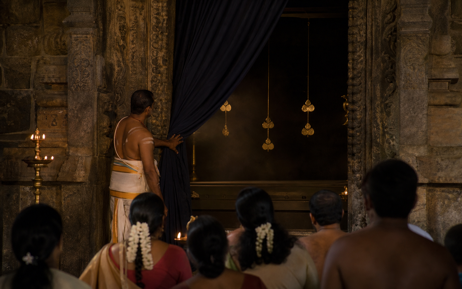 A temple curtain drawn aside before a dark, apparently empty sanctum recess decorated with golden bilva-leaf forms, as devotees observe from a distance.