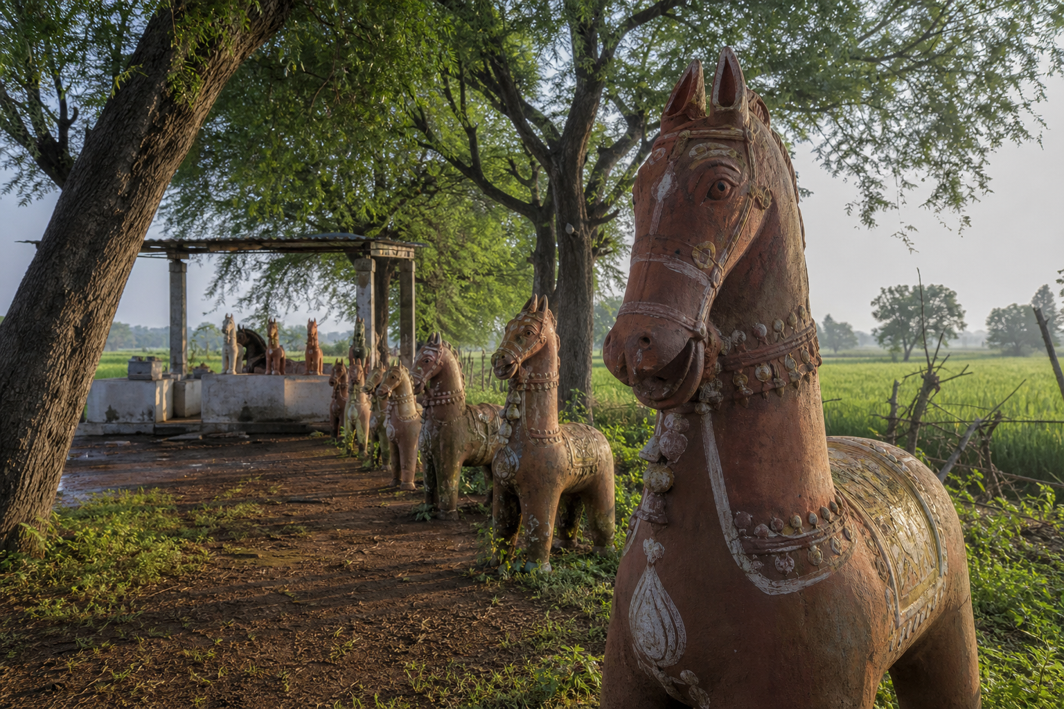 Rows of large terracotta horses stand among trees beside an Ayyanar shrine and cultivated fields in rural Tamil Nadu.