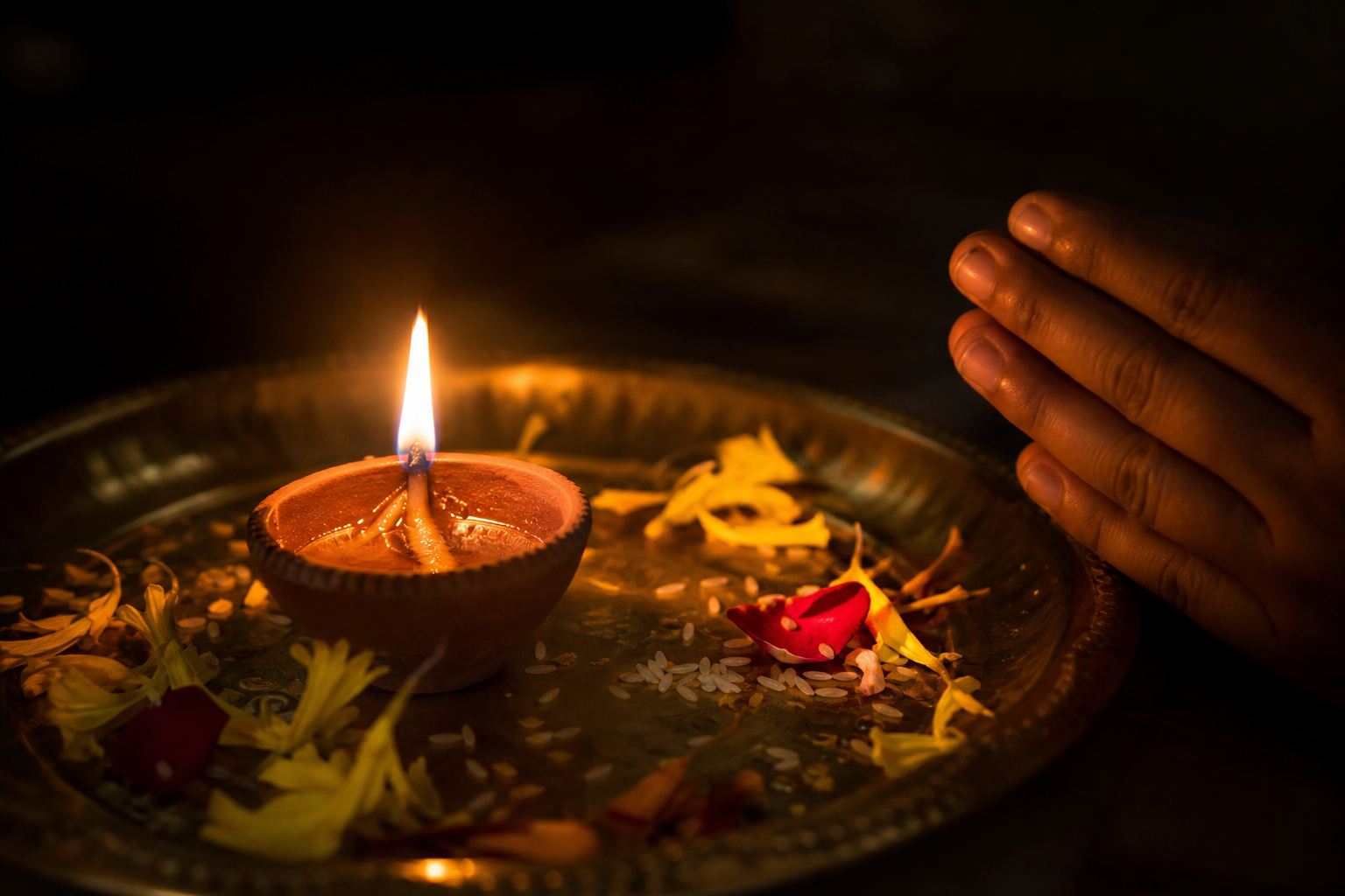 A clay oil lamp illuminates flower petals, rice grains, a brass plate, and folded hands in a dark room.