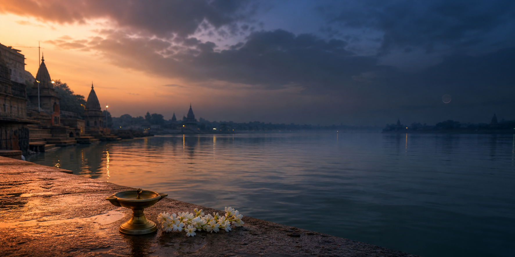 A brass oil lamp and white flowers beside a calm riverside ghat as daylight transitions into indigo twilight under a dark moon.