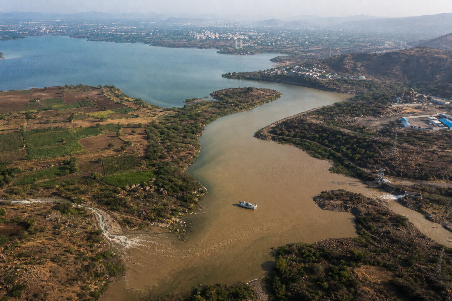 Aerial view of a reservoir and winding river with discolored inflows, farmland, distant development and a water-sampling boat.