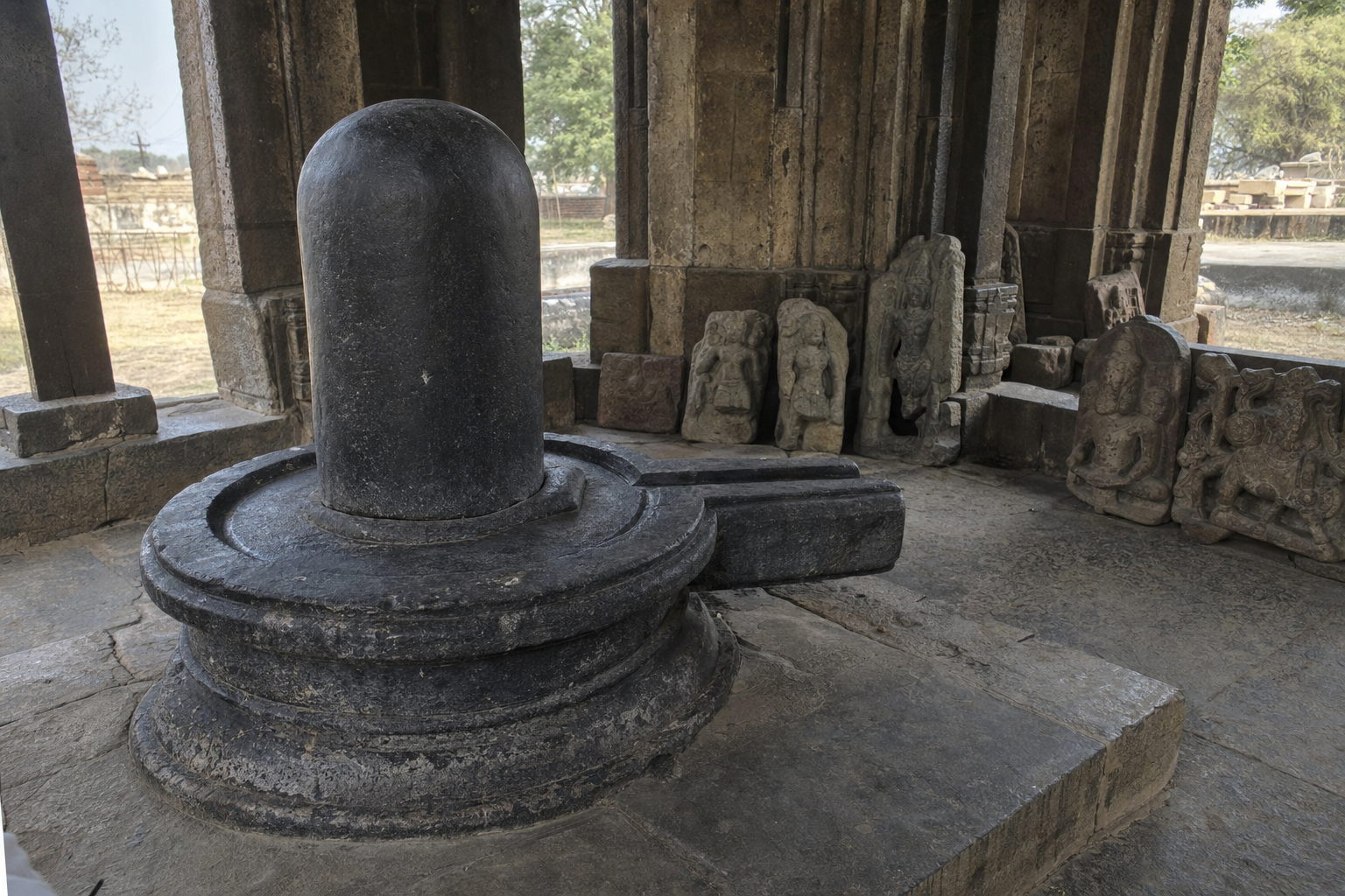 A large dark stone Shiva linga stands in a shaded temple space with weathered sculpture fragments positioned around the outer structure.