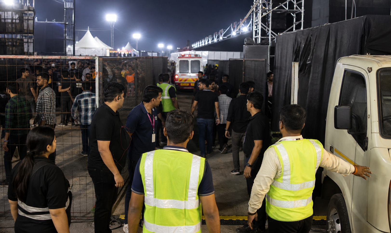Event staff inspect a narrow emergency corridor partly constrained by fencing, foot traffic, and a service vehicle near an ambulance bay.
