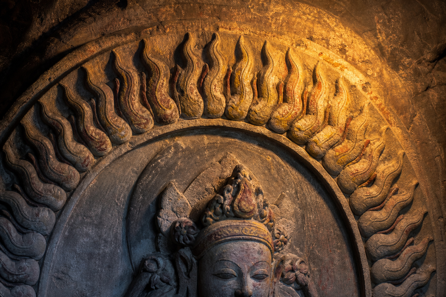 Close view of carved stone flames forming an aureole around part of a Jain goddess figure under warm lamp light.