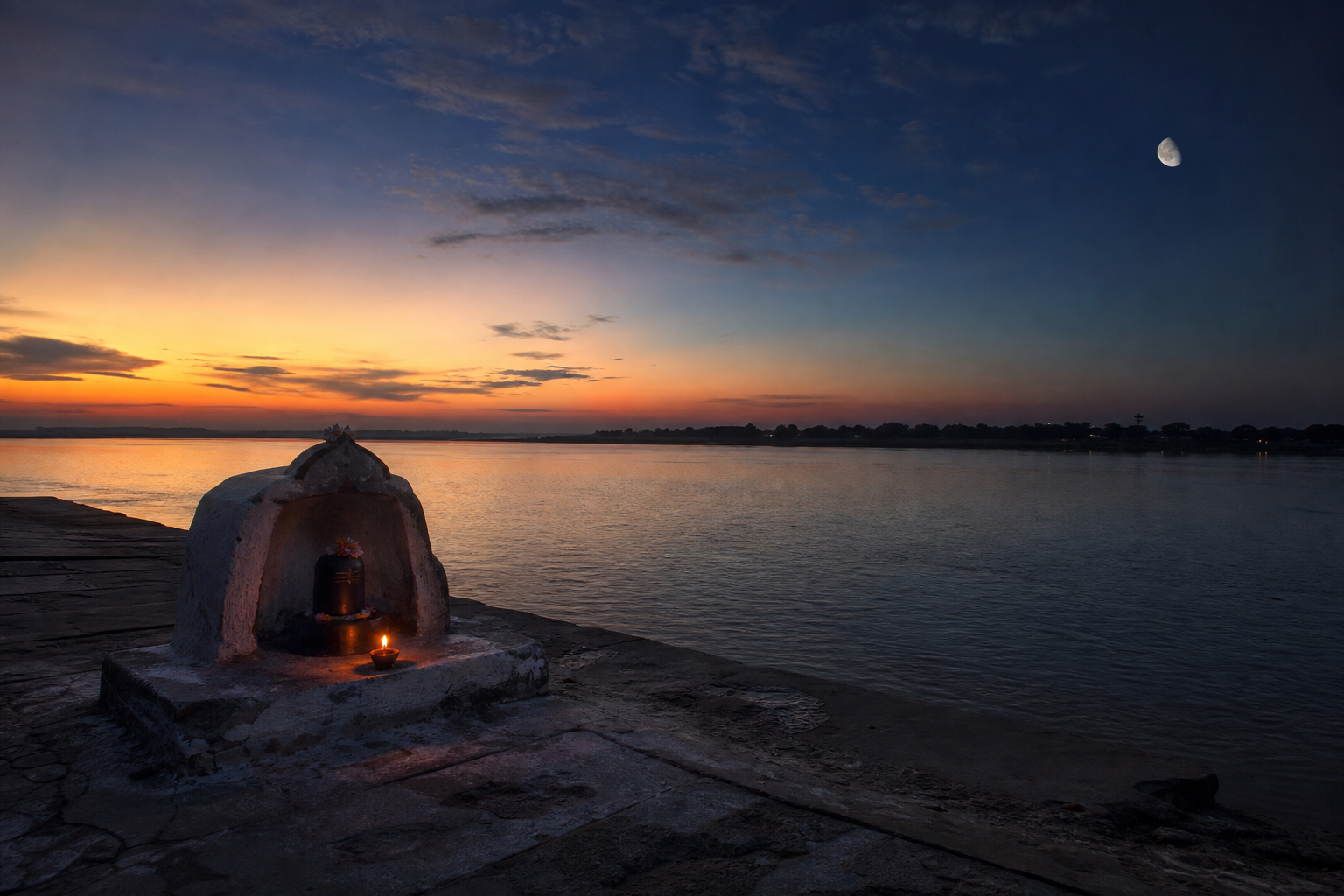 A small Shiva shrine beside a river at local sunset, with an oil lamp glowing and a waxing moon rising in the twilight sky.