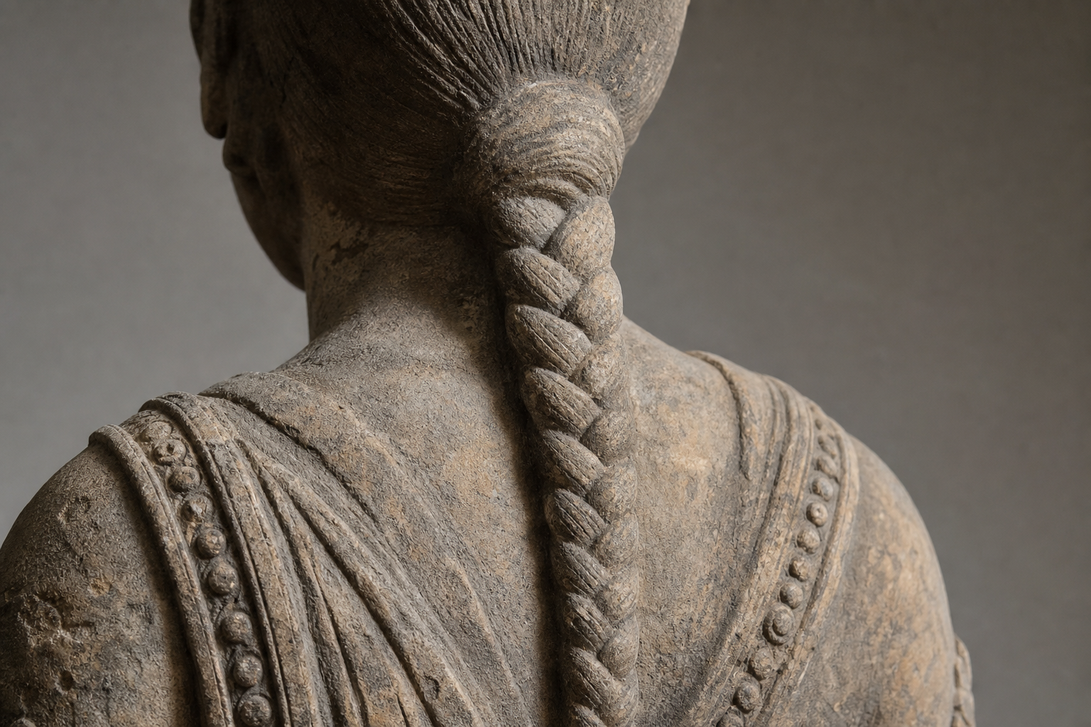 Close rear view of a weathered stone figure showing a single plait beginning at the nape and continuing down the back beside carved drapery and ornaments.
