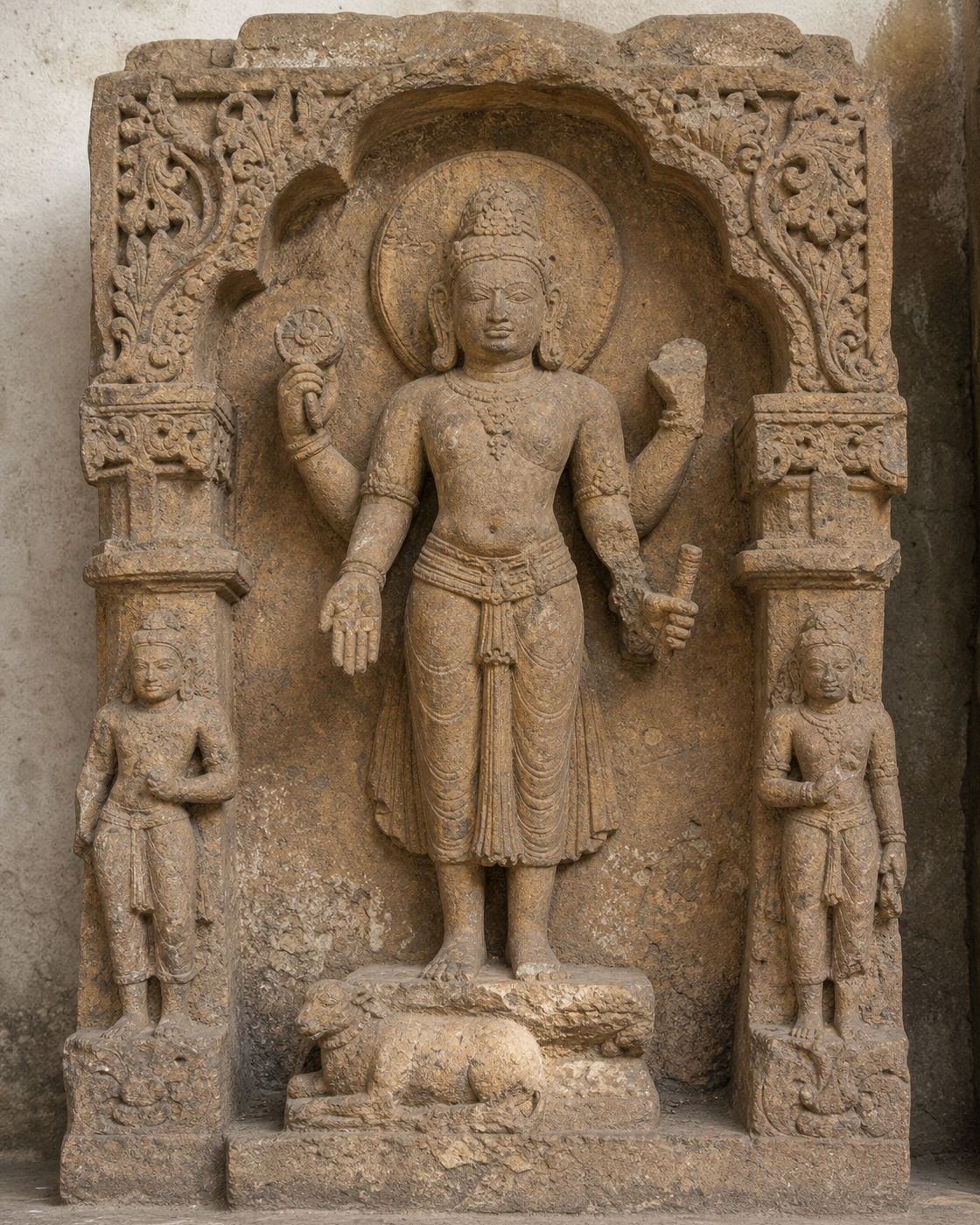 Weathered four-armed sandstone deity in a carved niche with an open palm, one surviving attribute, broken hands, two attendants, and an eroded pedestal animal.