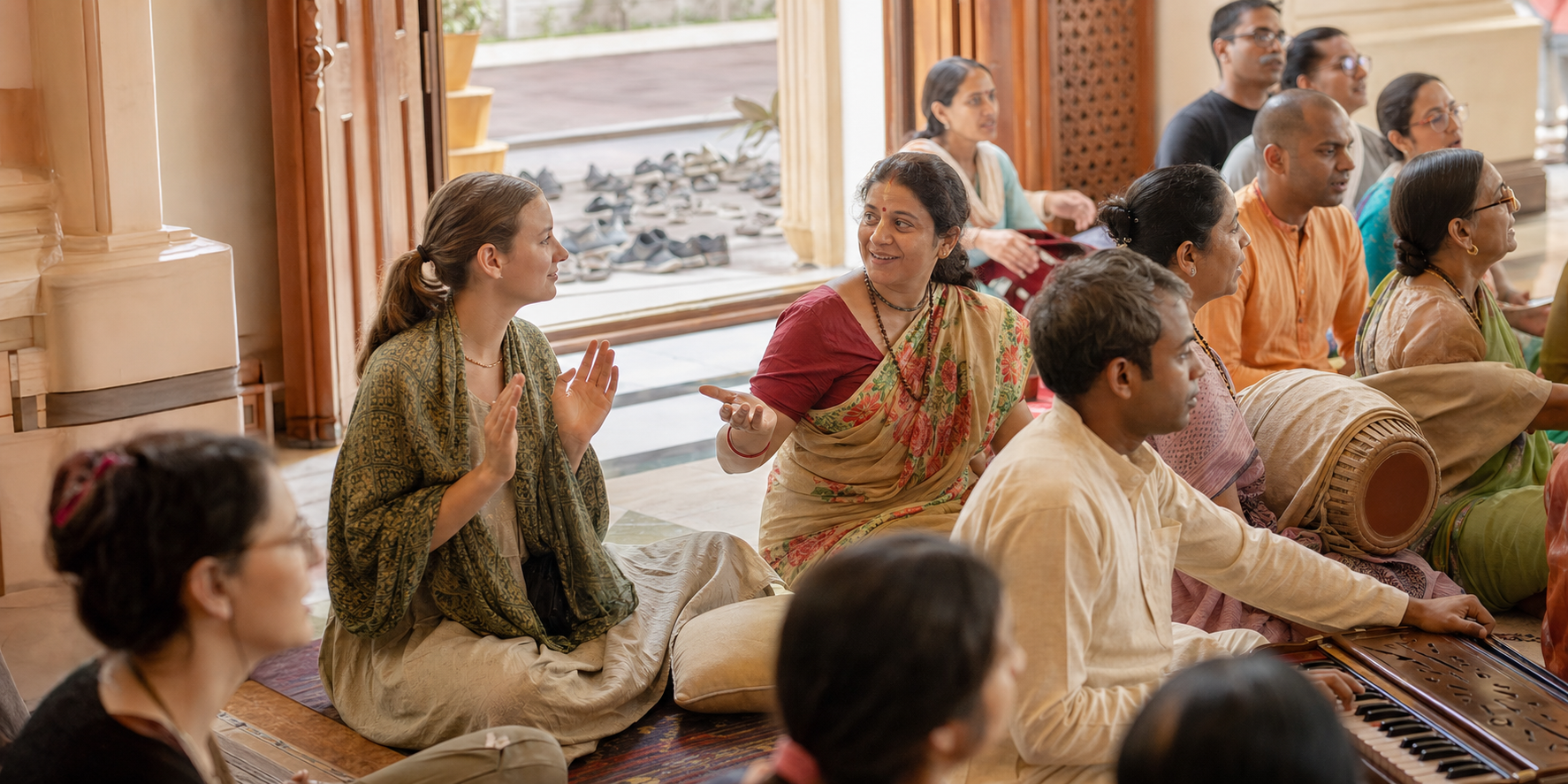 A newcomer joins a temple kirtan at the edge of the gathering while a nearby devotee offers a welcoming gesture.