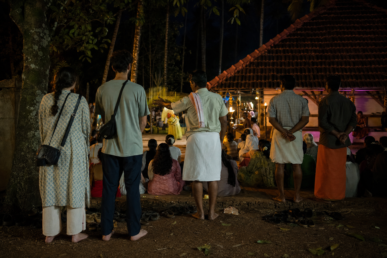 Two visitors stand quietly behind local families at a Bhuta Kola, with their footwear outside and a phone put away in a closed bag.