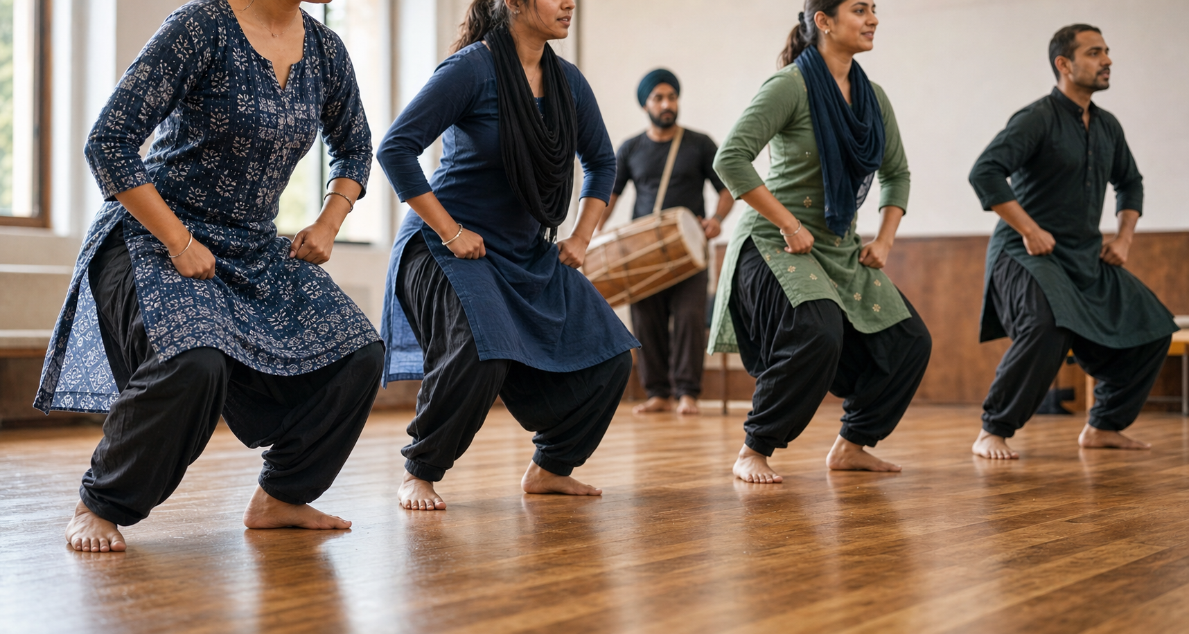 A small group of Punjabi folk dancers practices a synchronized grounded step while a dhol player stands in the background.