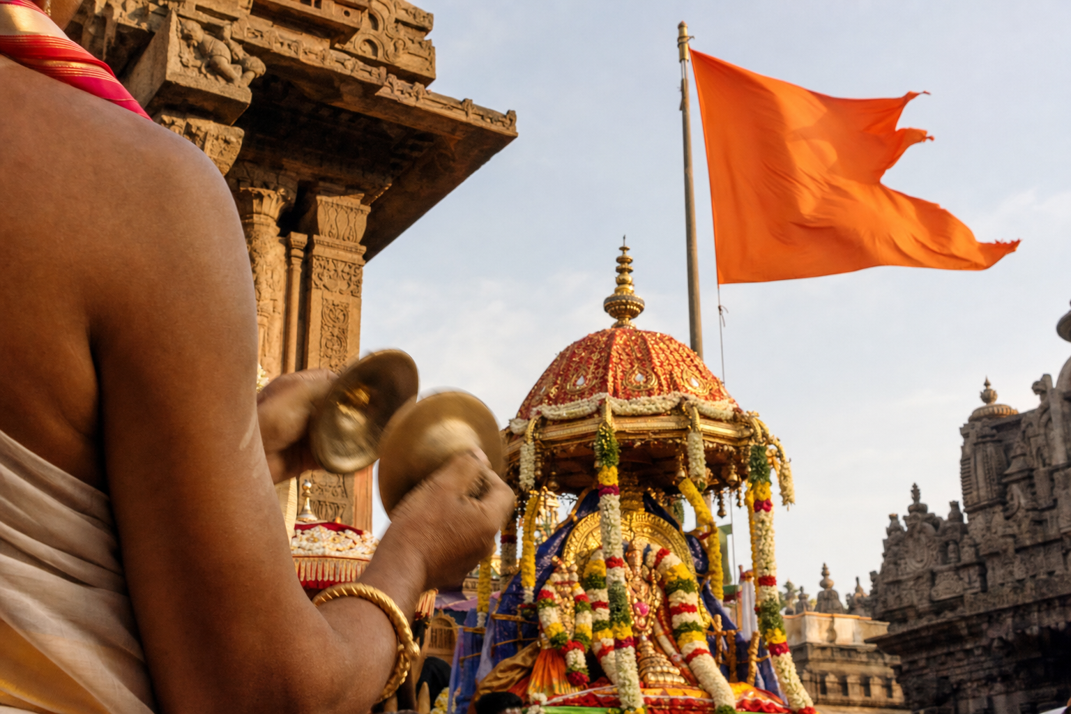 A saffron temple flag, bronze hand cymbals, flower garlands and a decorated shrine procession in a Hindu temple courtyard.