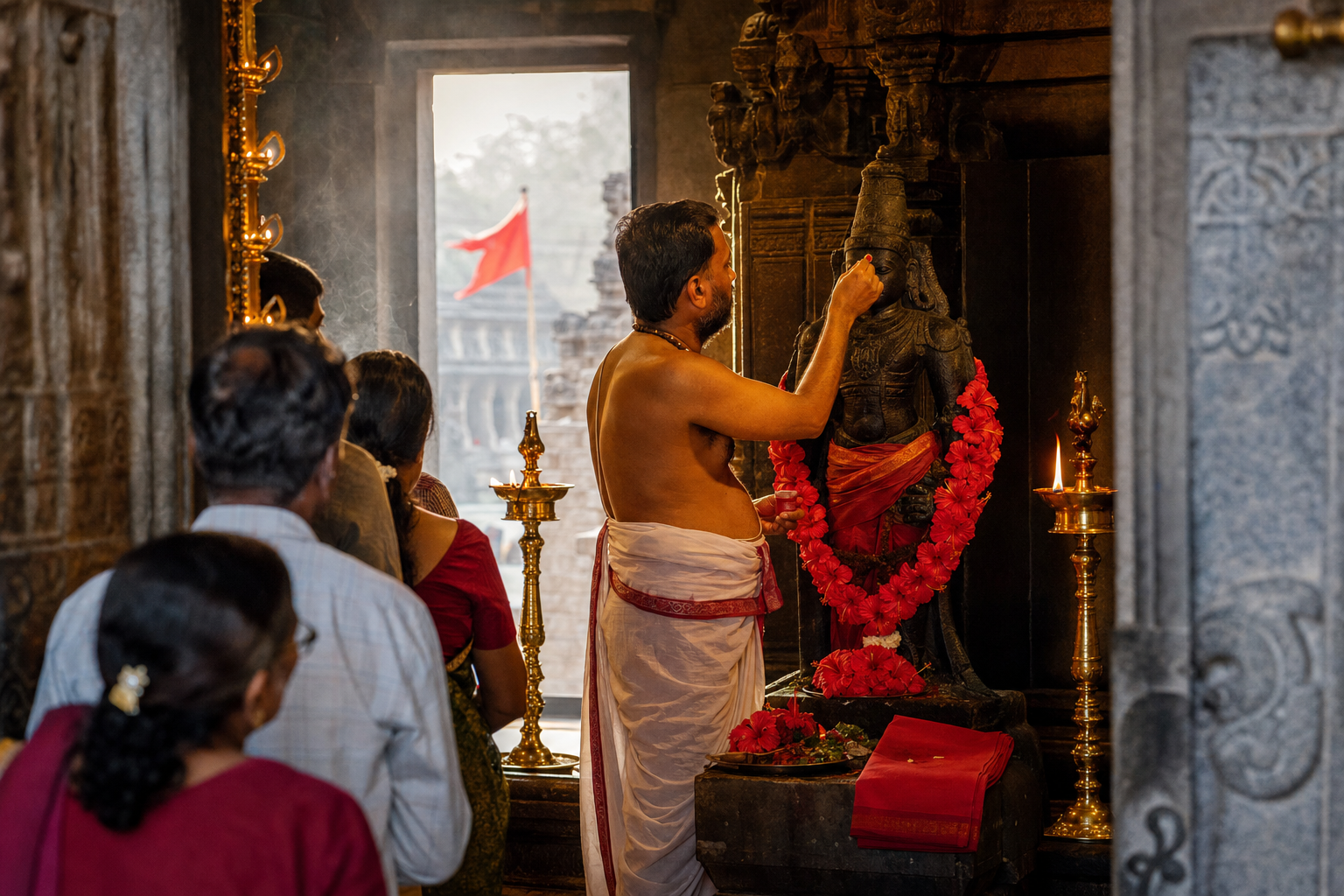 A priest applies red kumkum to a stone murti as devotees gather near a lamp-lit temple sanctum decorated with hibiscus and red cloth.