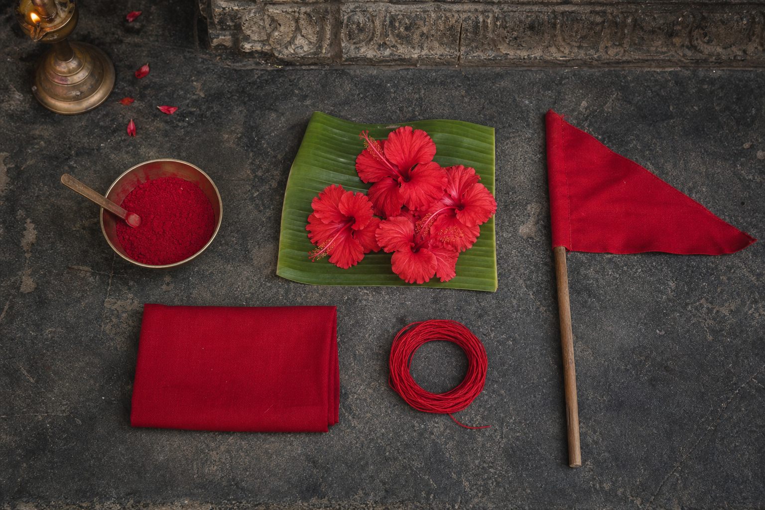A brass bowl of red kumkum, hibiscus flowers, folded red cloth, sacred thread, and a small triangular flag rest separately on a temple stone surface.