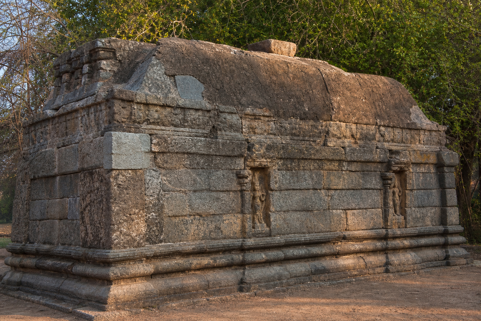 Low stone walls, sculptural niches, base mouldings, and the restrained roofline of the Keezha Kadambur shrine are seen in angled light.
