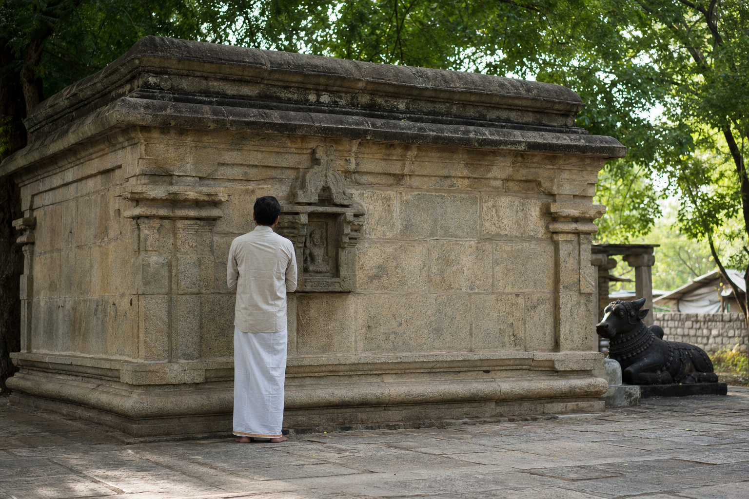 A devotee pauses to examine a weathered sculptural niche on the low stone wall of the Keezha Kadambur temple.