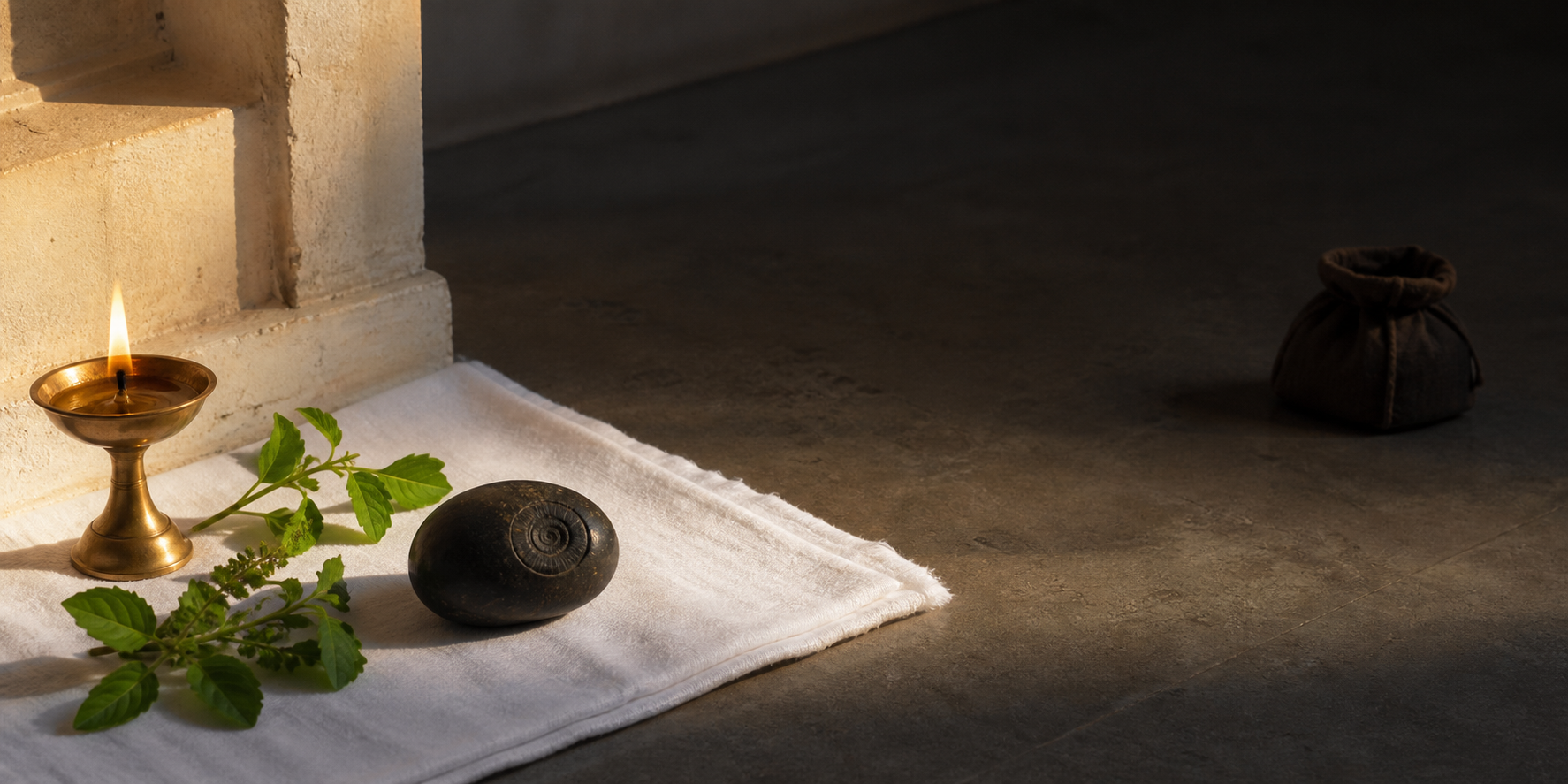 A Shaligram on a white cloth with Tulsi leaves and a lamp is separated by empty space from a closed coin pouch in shadow.