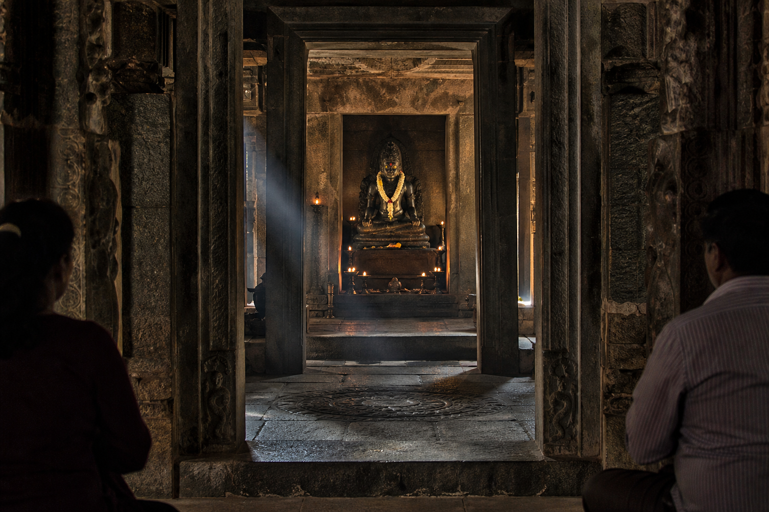 A flower-garlanded granite deity stands in a deep temple sanctum framed by carved stone, oil lamps, and incense haze.